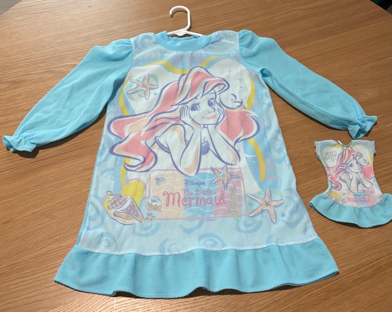 May include: A light blue nightgown featuring a graphic of Ariel from Disney's The Little Mermaid. The long-sleeved nightgown has a ruffled hem and cuffs. A matching doll dress is also present.