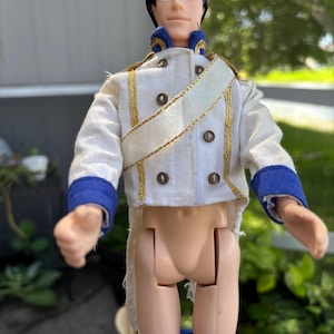 May include: A vintage doll of a prince wearing a white jacket with gold trim and blue accents. The doll has black hair and is wearing blue pants. The doll's jacket has gold buttons and a gold sash.