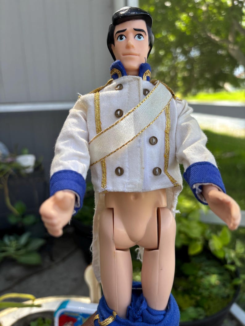 May include: A vintage doll of a prince wearing a white jacket with gold trim and blue accents. The doll has black hair and is wearing blue pants. The doll's jacket has gold buttons and a gold sash.