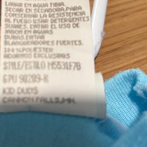 May include: Close-up of a white clothing tag with black text. The tag includes washing instructions and fabric content, with the text "100% POLIESTER" and "STYLE/ESTILO M5531F78". The tag is against a light blue fabric.