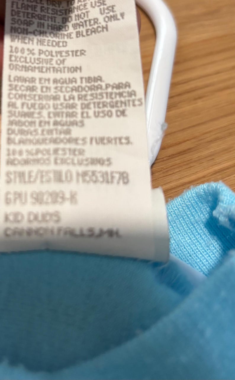 May include: Close-up of a white clothing tag with black text. The tag includes washing instructions and fabric content, with the text "100% POLIESTER" and "STYLE/ESTILO M5531F78". The tag is against a light blue fabric.
