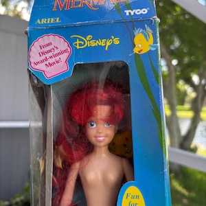 May include: A vintage Ariel doll from Disney's The Little Mermaid, in its original packaging. The doll has red hair, blue eyes, and a green mermaid tail. The box features the Disney's logo, the text "The Little Mermaid", and the text "Fun for water play, too!"