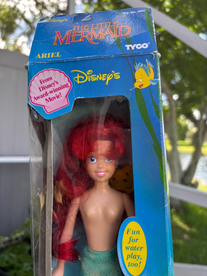 May include: A vintage Ariel doll from Disney's The Little Mermaid, in its original packaging. The doll has red hair, blue eyes, and a green mermaid tail. The box features the Disney's logo, the text "The Little Mermaid", and the text "Fun for water play, too!"