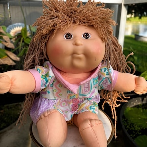 Puede incluir: Una muñeca con cara redonda, ojos marrones y cabello de lana marrón texturizado. La muñeca viste una camisa blanca con un patrón geométrico colorido, cuello rosa y dos botones rosas, combinada con pantalones cortos morados y piel color melocotón.
