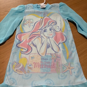 May include: A light blue nightgown featuring a graphic of Ariel from Disney's The Little Mermaid. The nightgown has long sleeves and a ruffled hem. The graphic includes Ariel, seashells, and starfish.