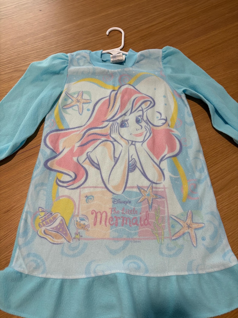 May include: A light blue nightgown featuring a graphic of Ariel from Disney's The Little Mermaid. The nightgown has long sleeves and a ruffled hem. The graphic includes Ariel, seashells, and starfish.