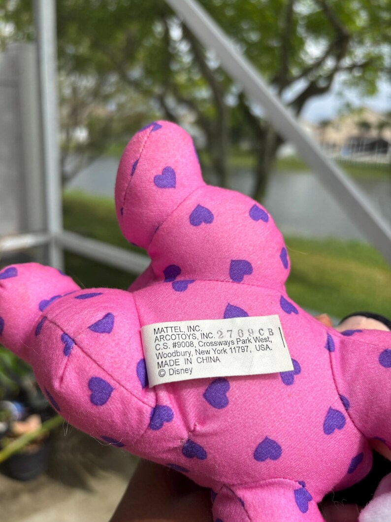 Pu&ograve; includere: Un peluche rosa con motivi a cuore viola. Il giocattolo ha un'etichetta che recita "MATTEL, INC. ARCOTOYS, INC. 2769 CB, MADE IN CHINA, &copy;Disney". &Egrave; un prodotto Disney.