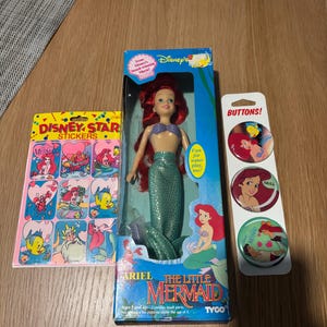 Pode incluir: Uma boneca da Ariel de A Pequena Sereia da Disney numa caixa azul, uma folha de autocolantes Disney Star com a Ariel e o Linguado, e um cart&atilde;o vertical com tr&ecirc;s bot&otilde;es circulares com a Ariel e o Linguado, todos com ilustra&ccedil;&otilde;es coloridas.