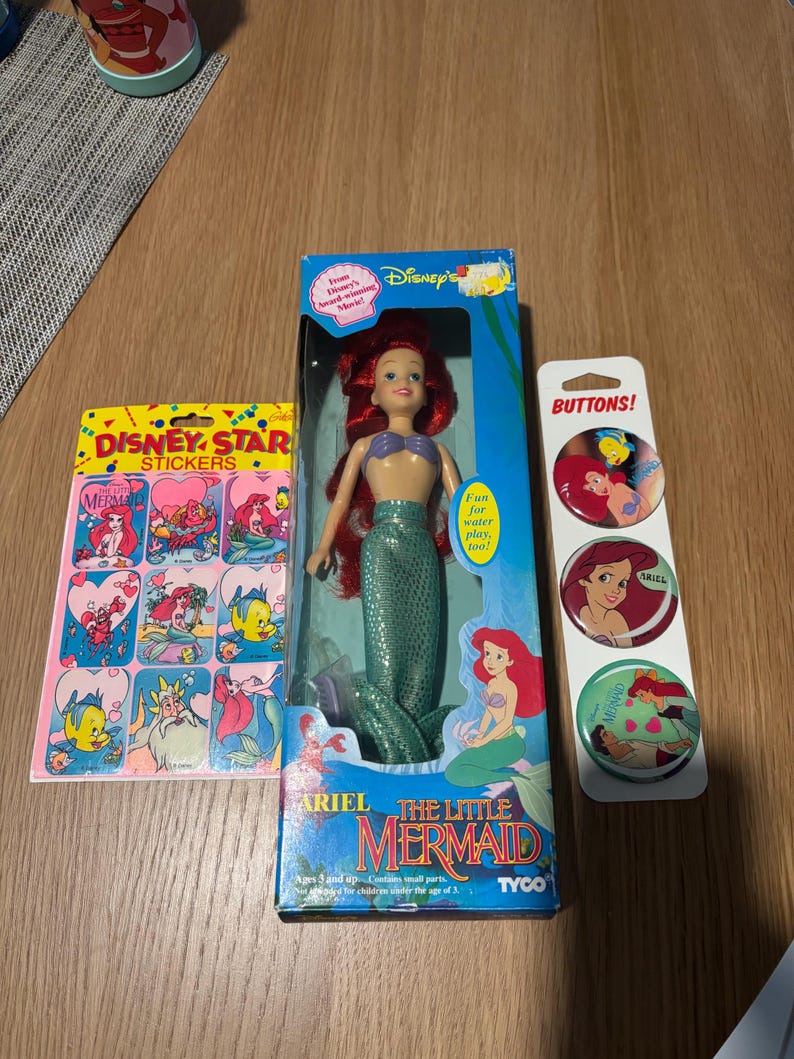 Pode incluir: Uma boneca da Ariel de A Pequena Sereia da Disney numa caixa azul, uma folha de autocolantes Disney Star com a Ariel e o Linguado, e um cart&atilde;o vertical com tr&ecirc;s bot&otilde;es circulares com a Ariel e o Linguado, todos com ilustra&ccedil;&otilde;es coloridas.
