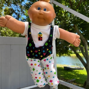 May include: A doll with a round head, light skin, blue eyes, and a small tuft of blonde hair. The doll wears a white short-sleeved shirt with a small geometric figure on the chest, paired with black and white overalls featuring colorful shapes.