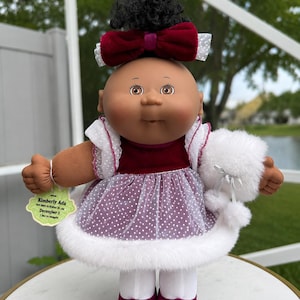 May include: A doll with dark skin and black curly hair styled in a top knot with a large burgundy velvet bow. The doll wears a burgundy velvet dress with a white polka-dot tulle overlay, white faux fur trim, white tights, and burgundy shoes.