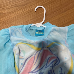 May include: A light blue long-sleeved shirt with a graphic of a mermaid. The shirt has a round neckline and is hanging on a white hanger. The mermaid graphic features pink, yellow, and blue colors.