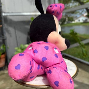 Pu&ograve; includere: Una bambola di peluche di Minnie Mouse, con testa nera, orecchie rosa e un fiocco rosa con pois viola. Il corpo della bambola &egrave; rosa con motivi a cuore viola. La bambola &egrave; seduta su un tavolino decorativo.