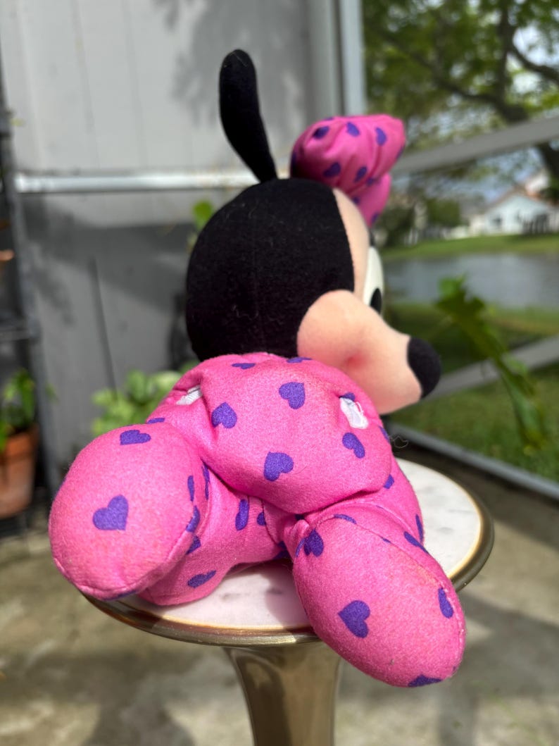 Pu&ograve; includere: Una bambola di peluche di Minnie Mouse, con testa nera, orecchie rosa e un fiocco rosa con pois viola. Il corpo della bambola &egrave; rosa con motivi a cuore viola. La bambola &egrave; seduta su un tavolino decorativo.
