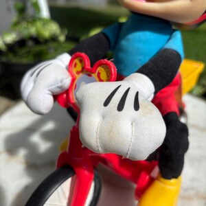 May include: A Mickey Mouse figurine riding a red and yellow tricycle. Mickey is wearing a blue shirt, red shorts, black gloves and shoes. The tricycle has black tires and a yellow basket.
