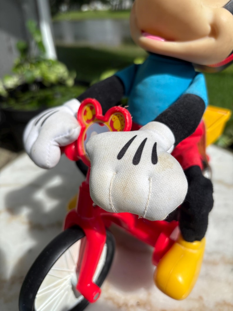 May include: A Mickey Mouse figurine riding a red and yellow tricycle. Mickey is wearing a blue shirt, red shorts, black gloves and shoes. The tricycle has black tires and a yellow basket.