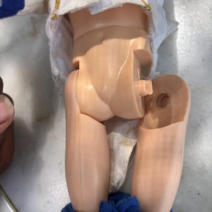 May include: A vintage doll with a white jacket with gold trim and blue accents. The doll's legs are detached, revealing the internal joint mechanism. The doll's blue pants are visible at the bottom of the image.