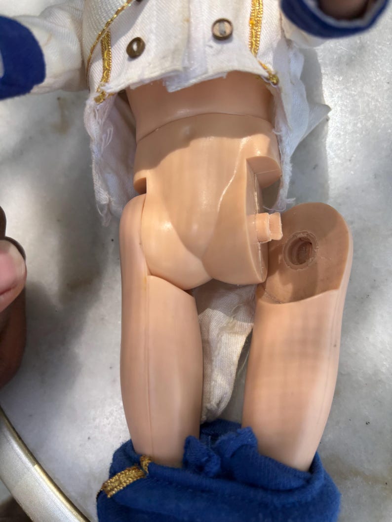 May include: A vintage doll with a white jacket with gold trim and blue accents. The doll's legs are detached, revealing the internal joint mechanism. The doll's blue pants are visible at the bottom of the image.