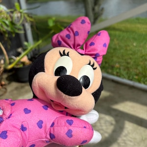 Pu&ograve; includere: Una bambola di peluche di Minnie Mouse con un motivo a cuori rosa e viola. La bambola ha la testa nera, grandi occhi e un fiocco rosa con cuori viola. La bambola indossa un completo rosa con cuori viola.