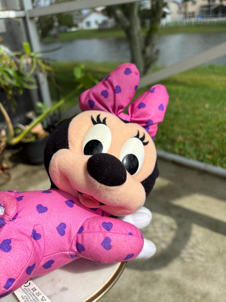 Pu&ograve; includere: Una bambola di peluche di Minnie Mouse con un motivo a cuori rosa e viola. La bambola ha la testa nera, grandi occhi e un fiocco rosa con cuori viola. La bambola indossa un completo rosa con cuori viola.