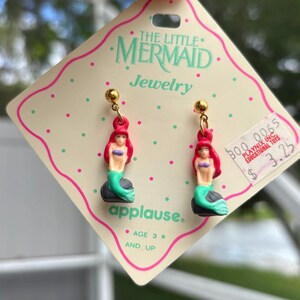 Op de afbeelding: Een paar bungelende oorbellen met De Kleine Zeemeermin. Elke oorbel heeft een goudkleurige knop en een miniatuurfiguur van Ariel met rood haar, een paarse top en een groene staart. De oorbellen zijn bevestigd aan een roze en witte kaart.