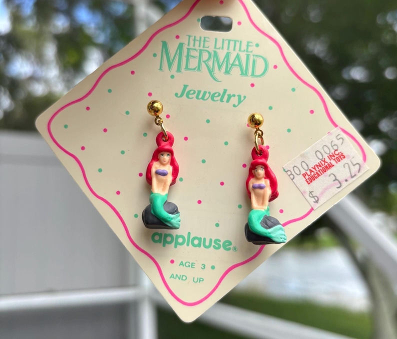 Op de afbeelding: Een paar bungelende oorbellen met De Kleine Zeemeermin. Elke oorbel heeft een goudkleurige knop en een miniatuurfiguur van Ariel met rood haar, een paarse top en een groene staart. De oorbellen zijn bevestigd aan een roze en witte kaart.
