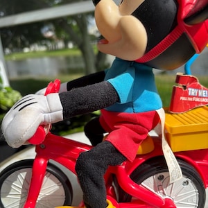 May include: A Mickey Mouse plush toy riding a red tricycle. Mickey is wearing a blue shirt, red shorts, yellow shoes, white gloves, and a red helmet. The tricycle has a yellow storage box on the back. The toy has the text "TRY ME!" on a tag.