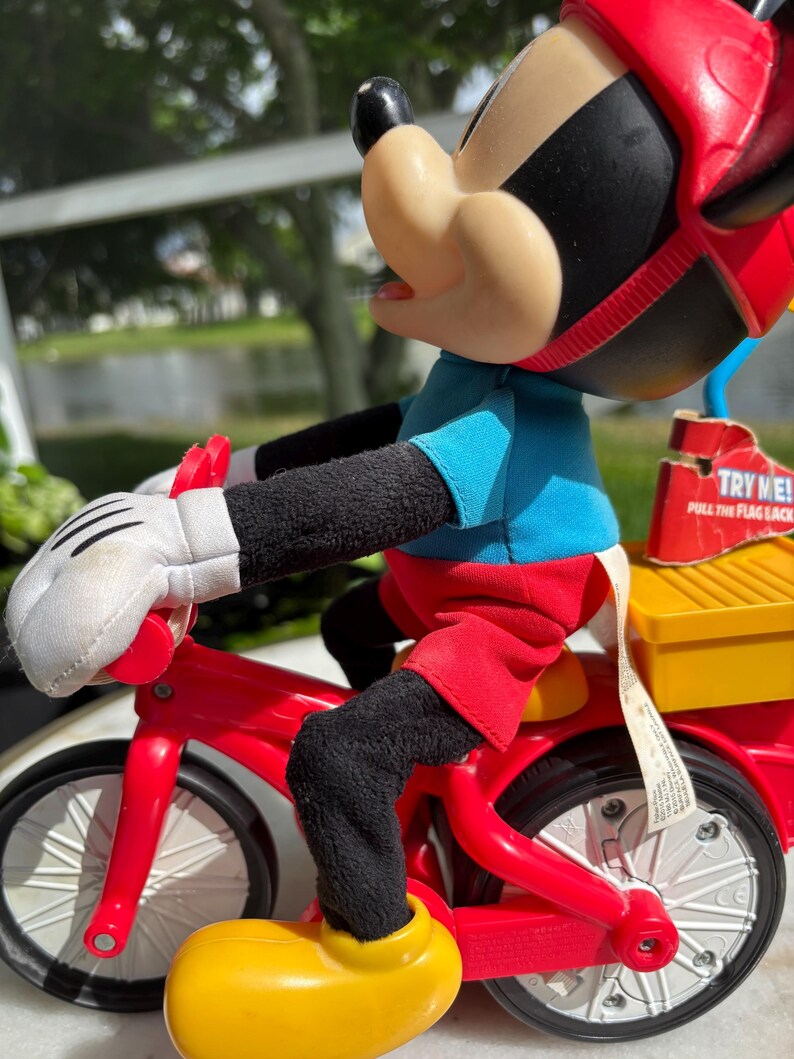 May include: A Mickey Mouse plush toy riding a red tricycle. Mickey is wearing a blue shirt, red shorts, yellow shoes, white gloves, and a red helmet. The tricycle has a yellow storage box on the back. The toy has the text "TRY ME!" on a tag.