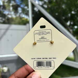 Op de afbeelding: Goudkleurige oorbellen met chirurgische stalen pinnen, gepresenteerd op een cr&egrave;mekleurig label. Het label bevat de tekst "EARRINGS" en een barcode. De oorbellen zijn van The Walt Disney Company.