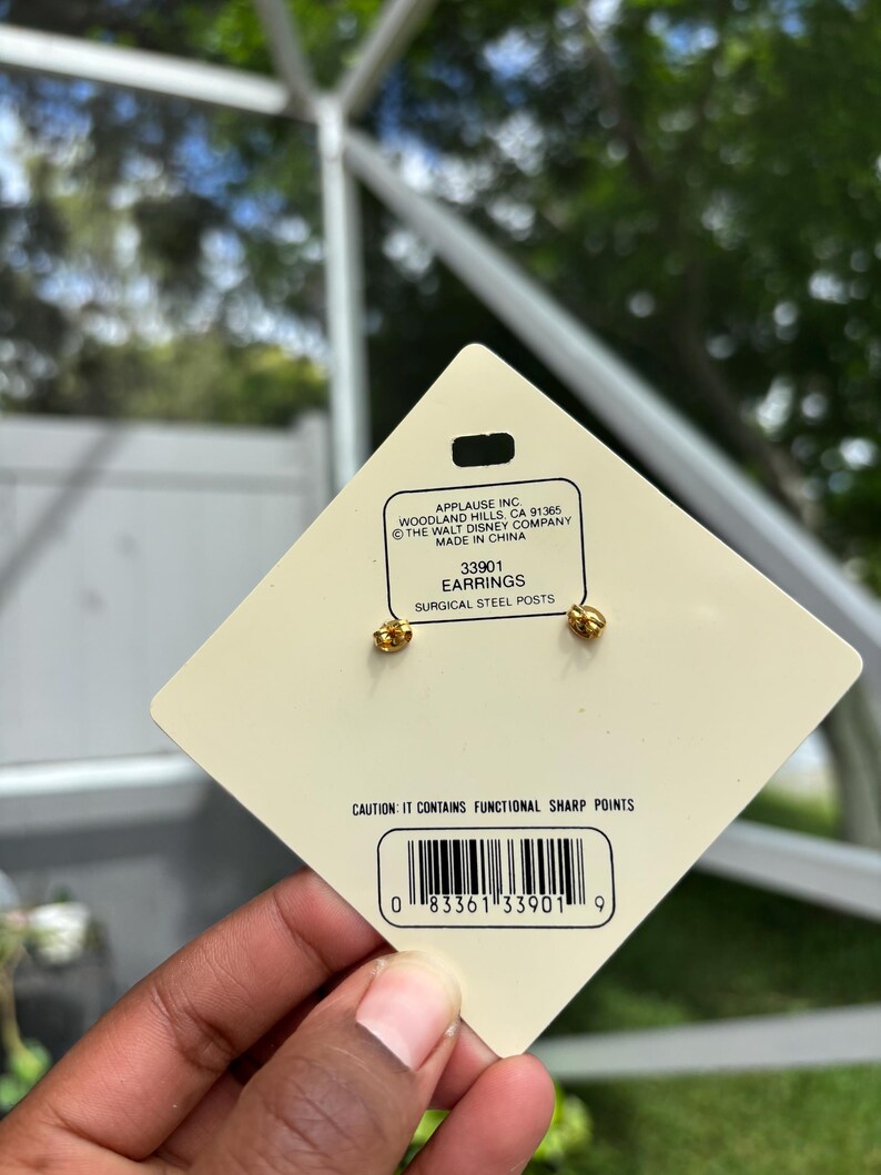 Op de afbeelding: Goudkleurige oorbellen met chirurgische stalen pinnen, gepresenteerd op een cr&egrave;mekleurig label. Het label bevat de tekst "EARRINGS" en een barcode. De oorbellen zijn van The Walt Disney Company.