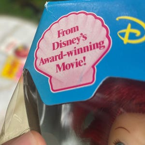 Pode incluir: Uma caixa de embalagem de cart&atilde;o azul com um gr&aacute;fico de concha rosa e o texto From Disney's Award-winning Movie! impresso a preto. Abaixo do texto, parte de uma boneca com cabelo vermelho vivo e olhos azuis &eacute; vis&iacute;vel no pl&aacute;stico.