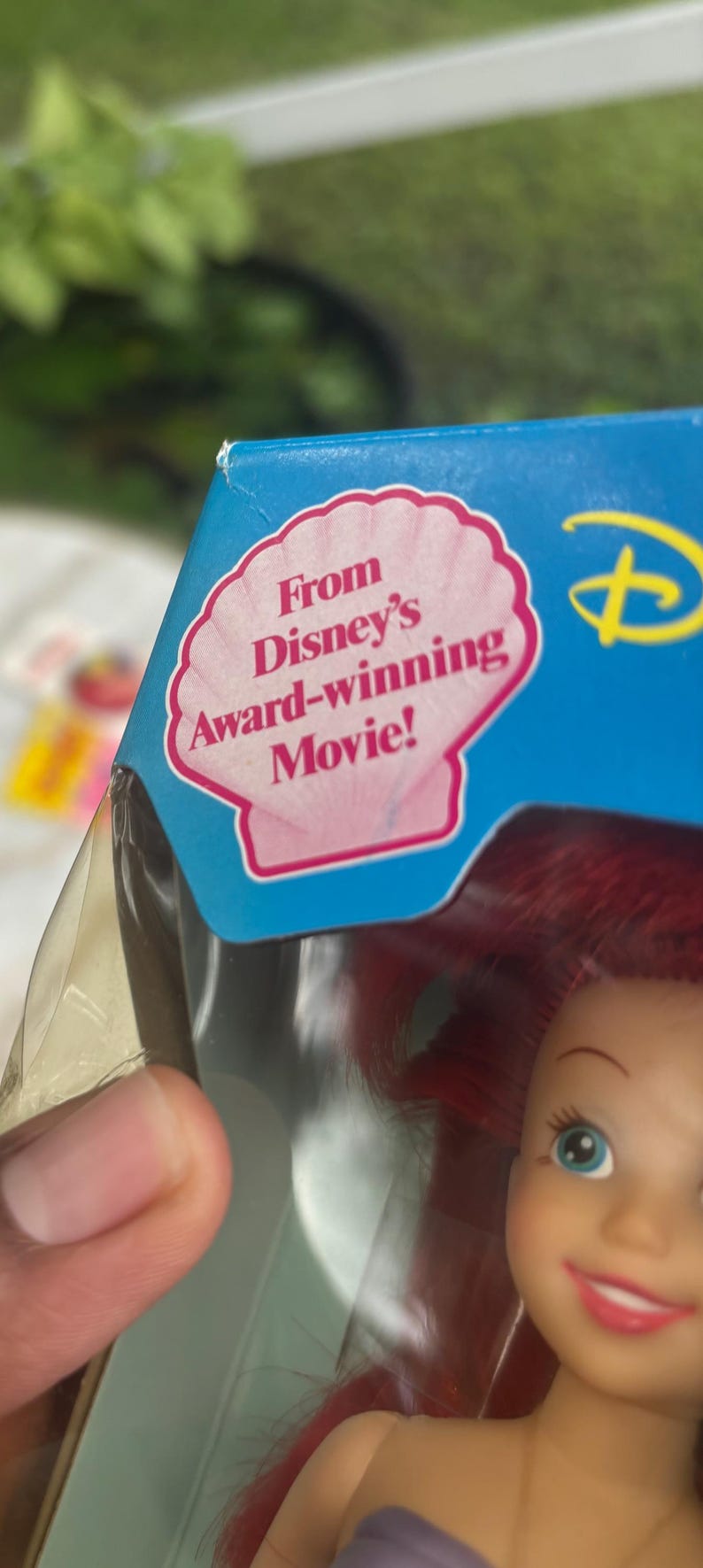 Pode incluir: Uma caixa de embalagem de cart&atilde;o azul com um gr&aacute;fico de concha rosa e o texto From Disney's Award-winning Movie! impresso a preto. Abaixo do texto, parte de uma boneca com cabelo vermelho vivo e olhos azuis &eacute; vis&iacute;vel no pl&aacute;stico.