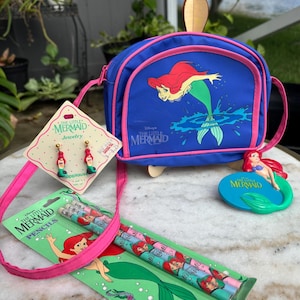 Op de afbeelding: Een verzameling "De Kleine Zeemeermin" merchandise. Inclusief een blauwe crossbodytas, oorbellen, potloden en een decoratief figuur. De tas en potloden tonen Ariel, de zeemeermin, in levendige kleuren.