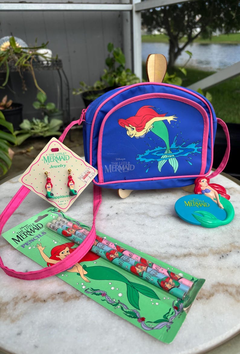 Op de afbeelding: Een verzameling "De Kleine Zeemeermin" merchandise. Inclusief een blauwe crossbodytas, oorbellen, potloden en een decoratief figuur. De tas en potloden tonen Ariel, de zeemeermin, in levendige kleuren.