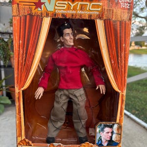 Może przedstawiać: Kolekcjonerska marionetka JC Chasez z *NSYNC, zapakowana w czerwono-pomarańczowe pudełko. Lalka ma na sobie czerwoną koszulę z długim rękawem i spodnie khaki. Pudełko zawiera logo Living Toyz i ostrzeżenie o małych częściach.