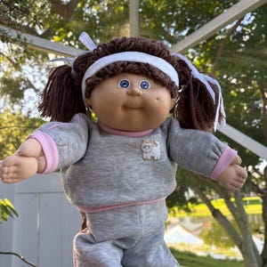 Könnte beinhalten: Eine Puppe mit braunem Wollhaar, das zu Zöpfen mit weißen Bändern und einem weißen Stirnband frisiert ist. Die Puppe hat blaue Augen, ein Grübchen an der linken Wange und trägt ein graues Langarmshirt mit rosa Saum und Katzenmotiv.
