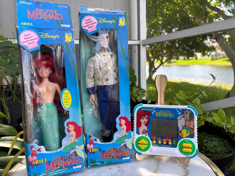 May include: Two vintage Disney's The Little Mermaid dolls in their original boxes, Ariel and Eric, along with an electronic handheld game. The Ariel doll has red hair and a green tail. The Eric doll is wearing a cream jacket and blue pants. The game features Ariel and other characters.