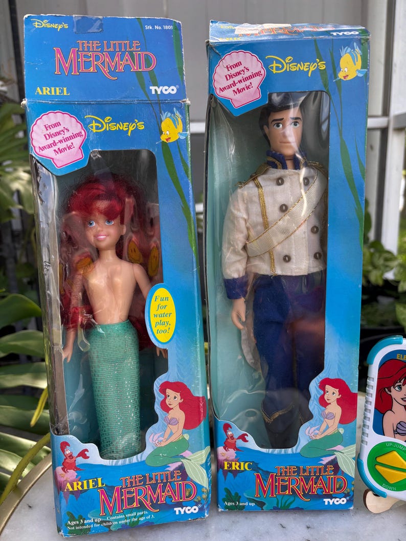 May include: Two vintage Disney's The Little Mermaid dolls in their original boxes. One doll is Ariel, with red hair and a green tail. The other is Prince Eric, in a white jacket and blue pants. The boxes feature Disney's branding and the text "From Disney's Award-winning Movie!"