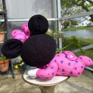 Pu&ograve; includere: Una bambola di peluche di Minnie Mouse in posizione reclinata. La bambola ha testa e orecchie nere, un fiocco rosa con pois viola e indossa un pigiama rosa con cuori viola. La bambola &egrave; appoggiata su un piccolo supporto decorativo.