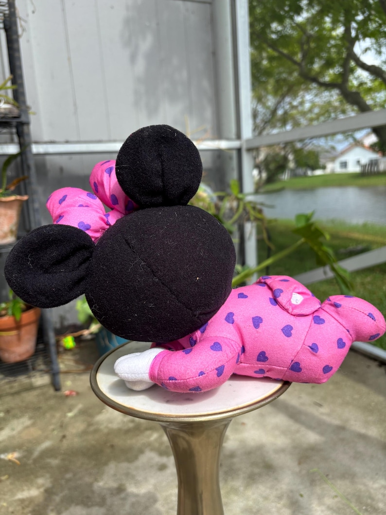 Pu&ograve; includere: Una bambola di peluche di Minnie Mouse in posizione reclinata. La bambola ha testa e orecchie nere, un fiocco rosa con pois viola e indossa un pigiama rosa con cuori viola. La bambola &egrave; appoggiata su un piccolo supporto decorativo.
