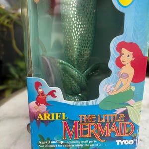 Pode incluir: Uma boneca Ariel A Pequena Sereia da Tyco na embalagem original. A caixa apresenta um gr&aacute;fico da Ariel e do Sebasti&atilde;o, com o texto Ariel The Little Mermaid, water play, too!, e Ages 3 and up. Contains small parts.