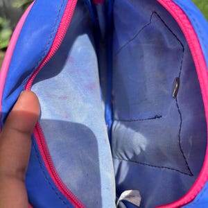 Op de afbeelding: Open blauwe en roze etui met ritssluiting. De etui heeft een blauwe buitenkant met een roze rits en rand. De binnenkant is lichtblauw met een klein vakje. De etui is open en onthult de binnenkant.