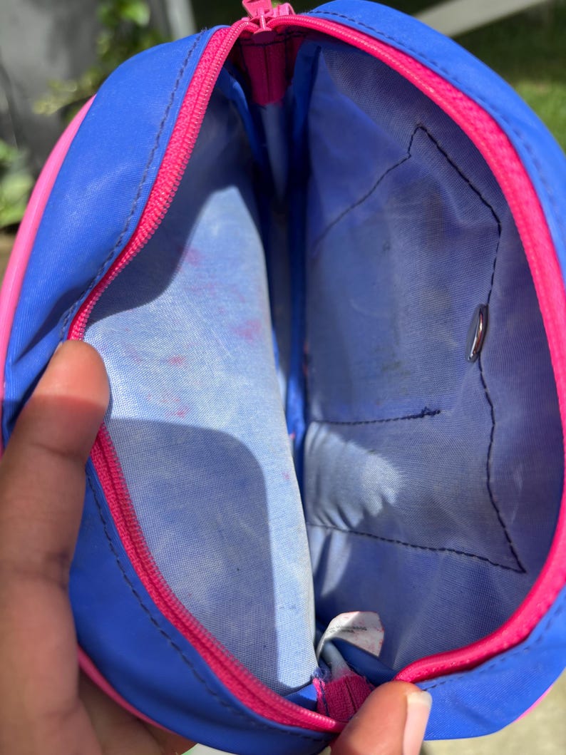 Op de afbeelding: Open blauwe en roze etui met ritssluiting. De etui heeft een blauwe buitenkant met een roze rits en rand. De binnenkant is lichtblauw met een klein vakje. De etui is open en onthult de binnenkant.