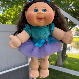 Könnte beinhalten: Eine Puppe mit dunkelbraunem Haar, blauen Augen und rosa Wangen, bekleidet mit einem glitzernden türkisfarbenen Oberteil und einem violetten Tüllrock. Eine türkisfarbene Schleife ist im Haar befestigt und ein türkisfarbenes Band mit einer kleinen violetten Blume ist um die Taille gebunden.