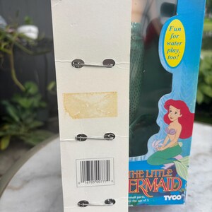 Pode incluir: Um cart&atilde;o de suporte de papel&atilde;o branco vertical com tr&ecirc;s alfinetes de metal presos, apresentando um c&oacute;digo de barras e texto. Ao lado, uma caixa azul para uma boneca da Pequena Sereia, com uma ilustra&ccedil;&atilde;o e o texto Fun for water play, too!