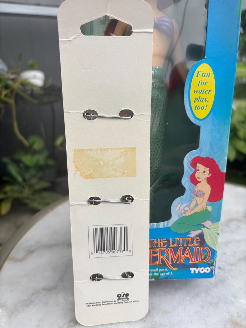 Pode incluir: Um cart&atilde;o de suporte de papel&atilde;o branco vertical com tr&ecirc;s alfinetes de metal presos, apresentando um c&oacute;digo de barras e texto. Ao lado, uma caixa azul para uma boneca da Pequena Sereia, com uma ilustra&ccedil;&atilde;o e o texto Fun for water play, too!