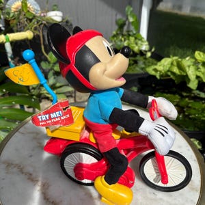 May include: A vintage Mickey Mouse toy figurine riding a red tricycle. Mickey wears a red helmet, blue shirt, red shorts, and yellow shoes. The tricycle has a yellow box with the text "TRY ME! PULL THE FLAG BACK". A blue flag is attached.