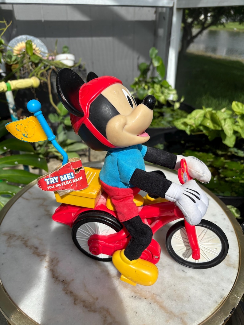 May include: A vintage Mickey Mouse toy figurine riding a red tricycle. Mickey wears a red helmet, blue shirt, red shorts, and yellow shoes. The tricycle has a yellow box with the text "TRY ME! PULL THE FLAG BACK". A blue flag is attached.