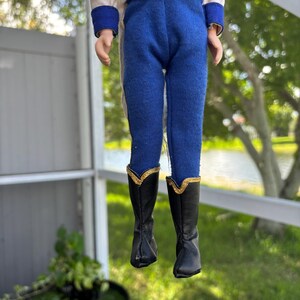 May include: A doll wearing a white jacket with gold trim and blue pants. The doll's black boots have gold accents. The doll is suspended in front of a blurred outdoor background.