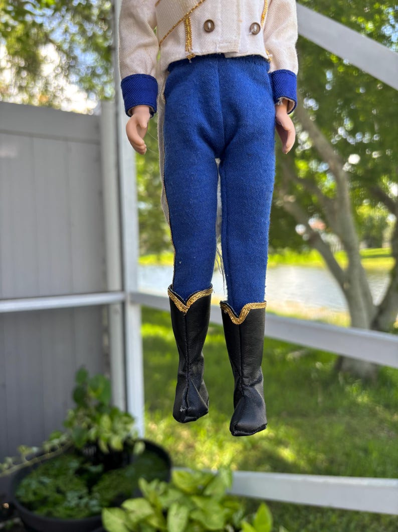 May include: A doll wearing a white jacket with gold trim and blue pants. The doll's black boots have gold accents. The doll is suspended in front of a blurred outdoor background.